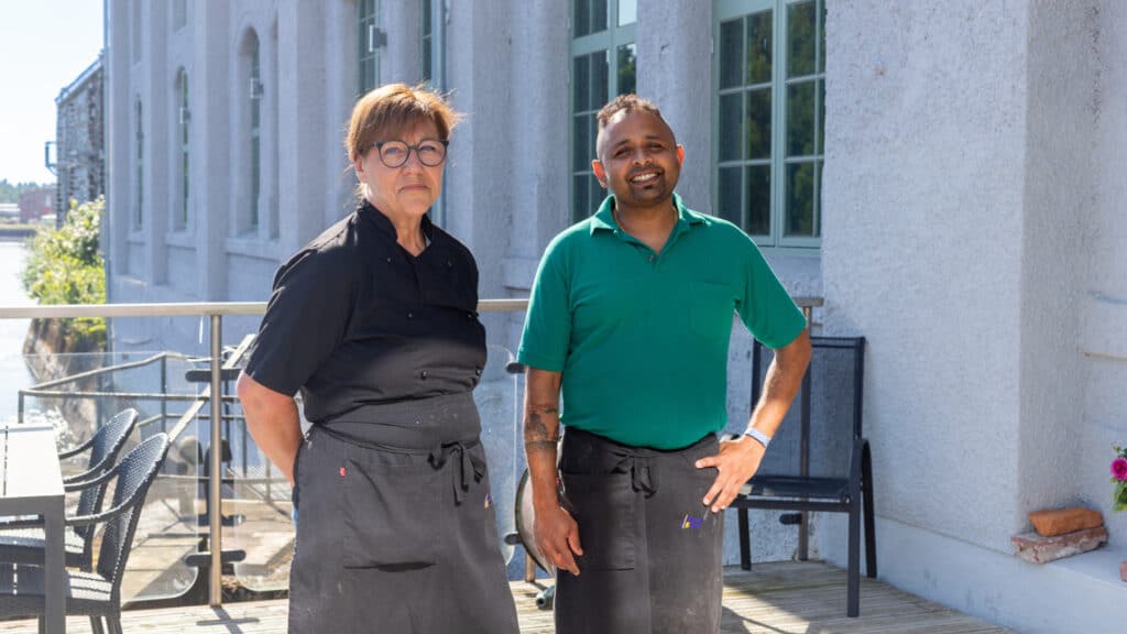 En kvinna och en man står ute på en uteservering med grå husfasad i bakgrunden.