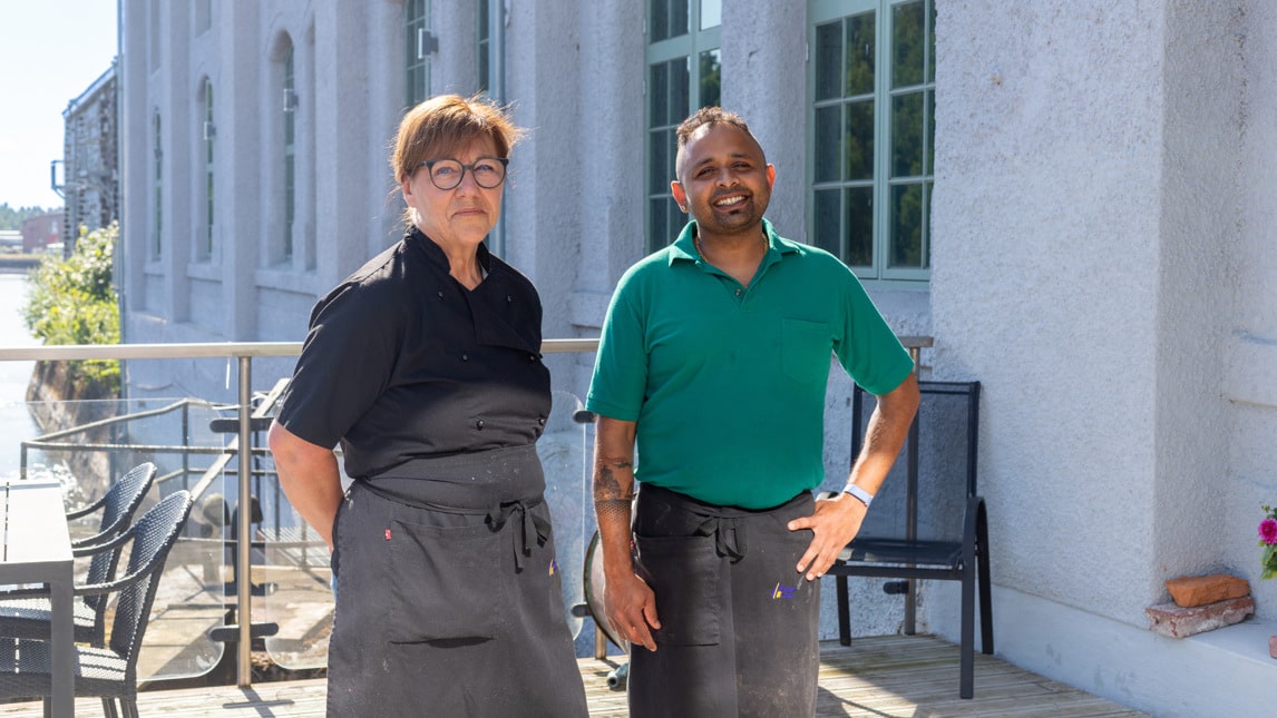 En kvinna och en man står ute på en uteservering med grå husfasad i bakgrunden.
